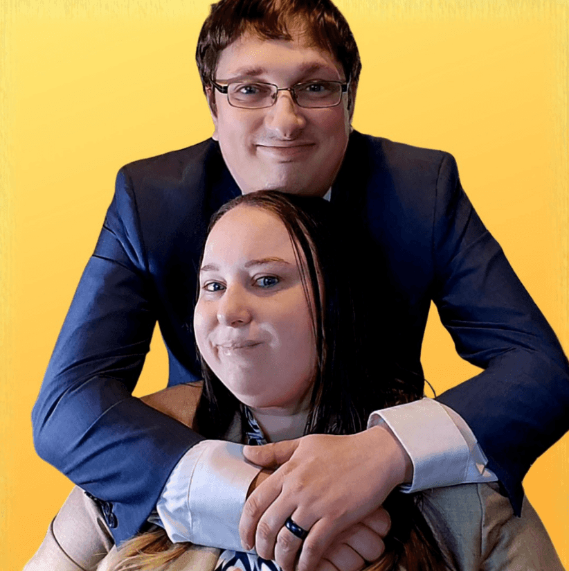 Smiling man in a blue suit hugs a seated woman from behind in front of a yellow background.