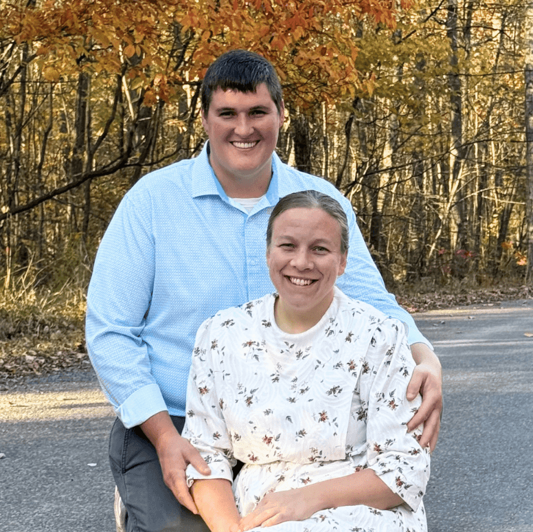 Smiling couple posing outdoors in autumn, the man standing behind the seated woman with his arm around her shoulders.