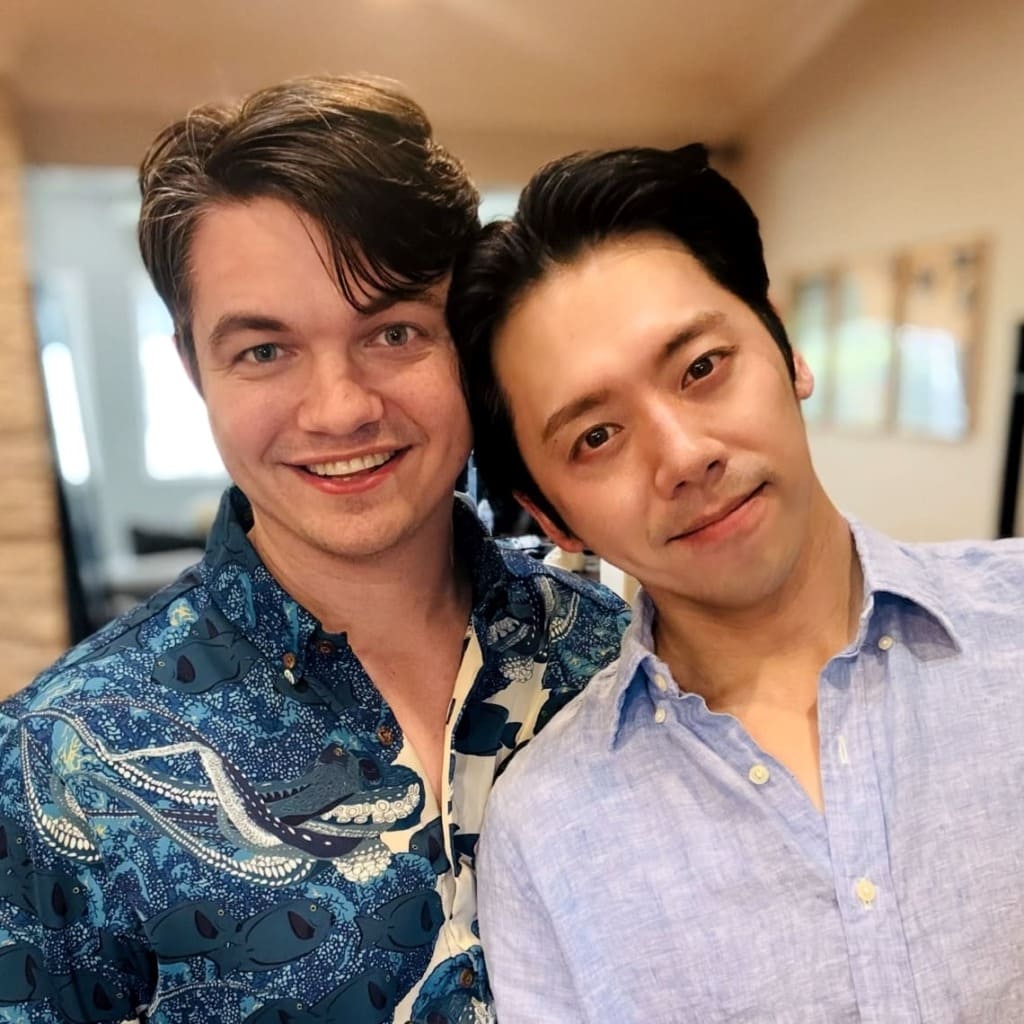 Two men take a friendly selfie indoors, smiling at the camera; one wears a blue patterned shirt and the other a light blue button-down.