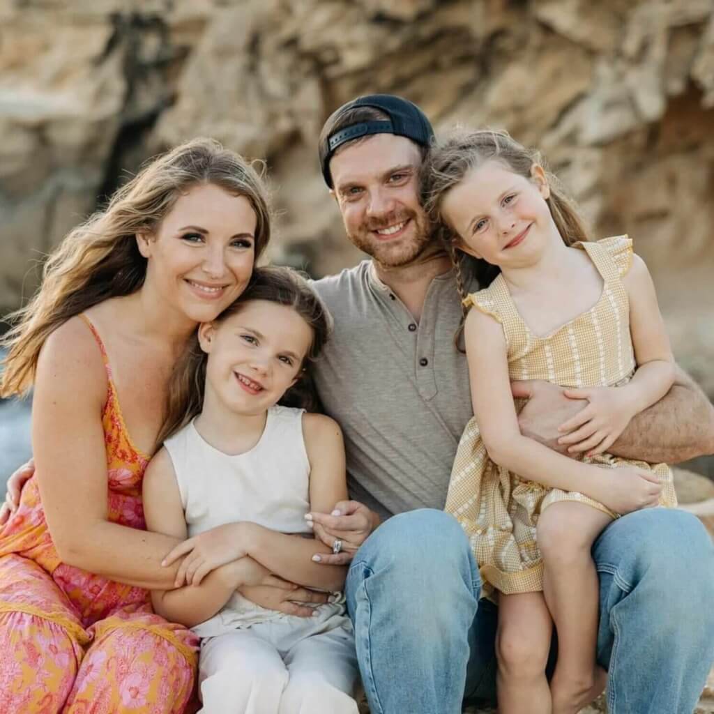 Smiling family of four posing on a rocky beach: mother in pink dress, father in gray shirt and cap, two daughters in light outfits.