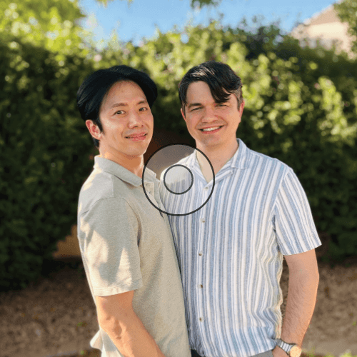 Two men posing outdoors, smiling at the camera with green hedges in the background.