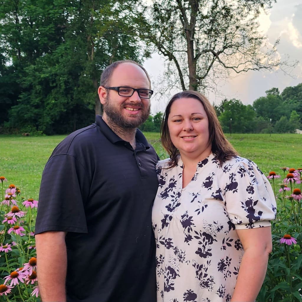 Smiling couple stands in a flower garden with pink coneflowers and green trees in the background.